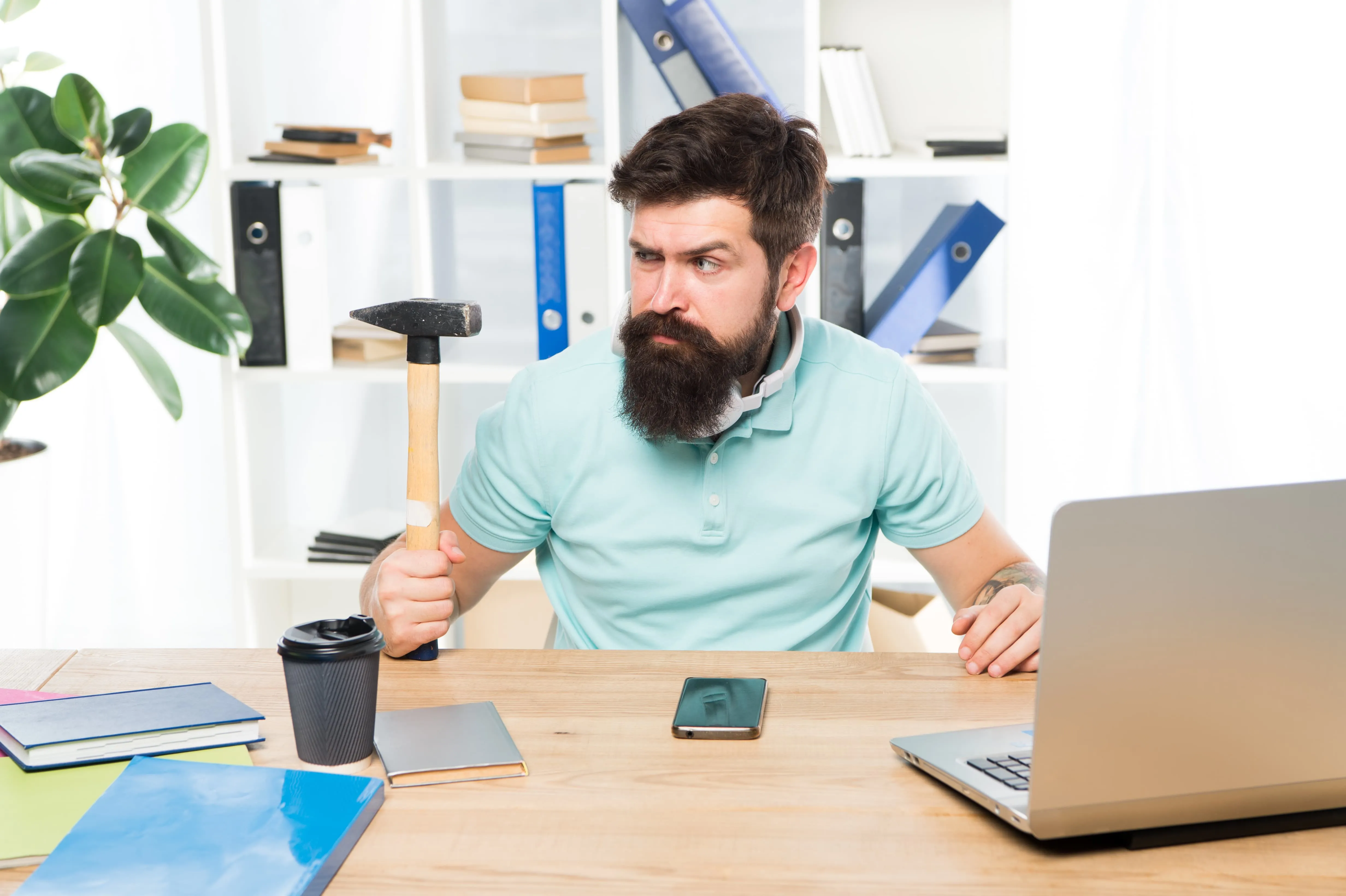 A seated bearded man holding a hammer in his left hand while looking pensively at it, suggesting that he is tempted to use it to break the computer that is in front of him